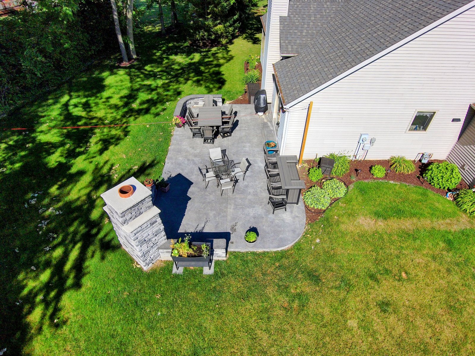 Concrete patios in Pierceton. Aerial view of a backyard concrete patio attached to a light-colored house, featuring multiple seating areas, a stone outdoor fireplace, and landscaped borders with grass and shrubs. Concrete patios in Ossian. Concrete contractor in Middlebury
