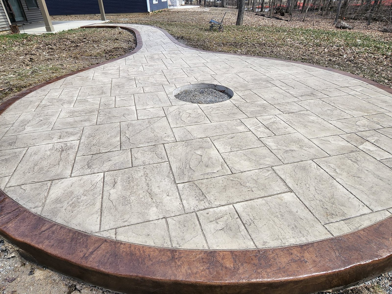 Decorative stamped concrete patio with a textured, light stone-like finish, featuring a circular gravel fire pit in the center and a curved dark-stained border, leading to a wooded backyard. Concrete patios in Huntington.