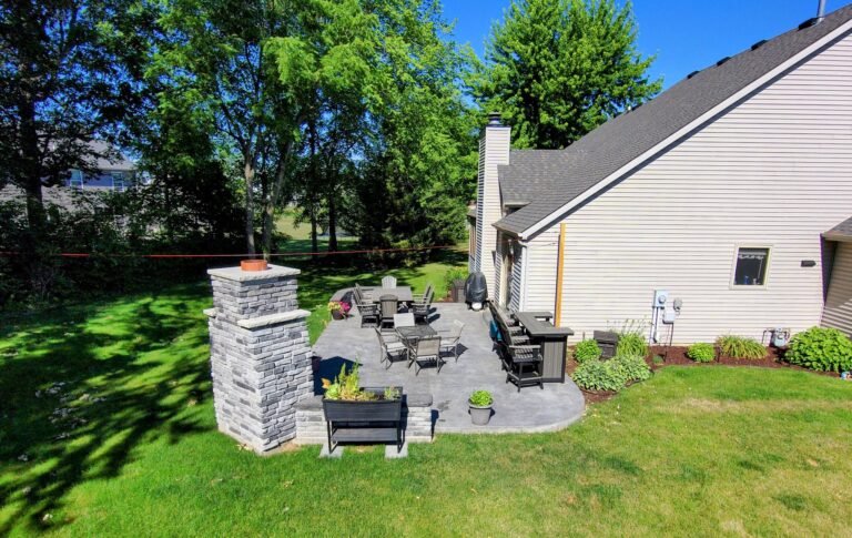 Backyard stamped concrete patio with outdoor dining, seating, and a built-in bar area surrounded by mature trees and greenery.