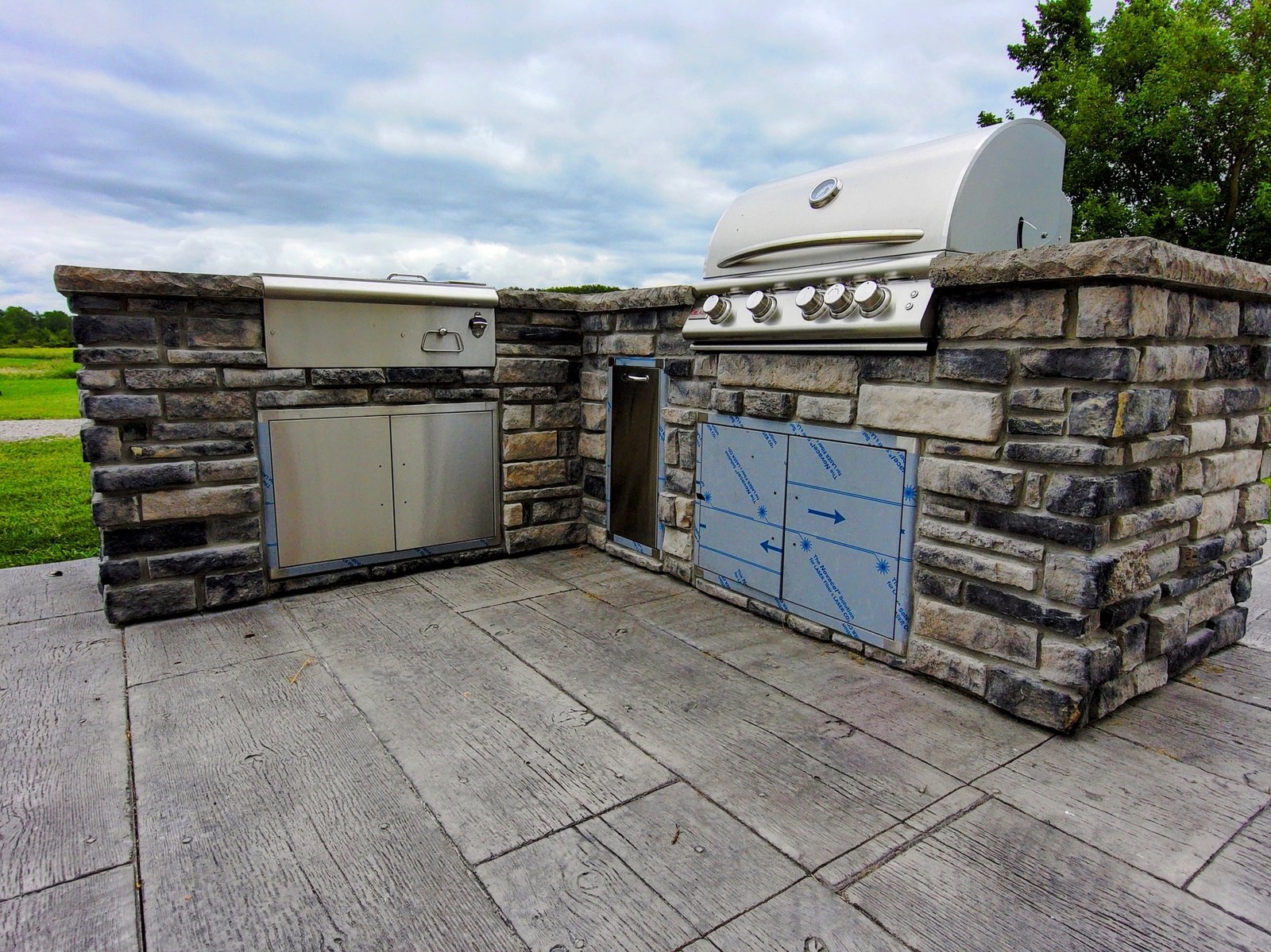 Outdoor kitchen built with stacked stone walls, featuring a built-in stainless steel grill, storage cabinets, and a concrete patio surface, set against a grassy landscape. Stamped concrete in Butler. Concrete patios in Defiance