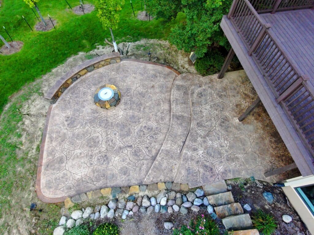 Stamped Concrete in Fort Wayne. Aerial view of a multi-level stamped concrete patio with natural stone texture and curved edges, featuring a built-in fire pit, accent borders, and stone stair pathway, seamlessly integrated into a wooded backyard landscape.