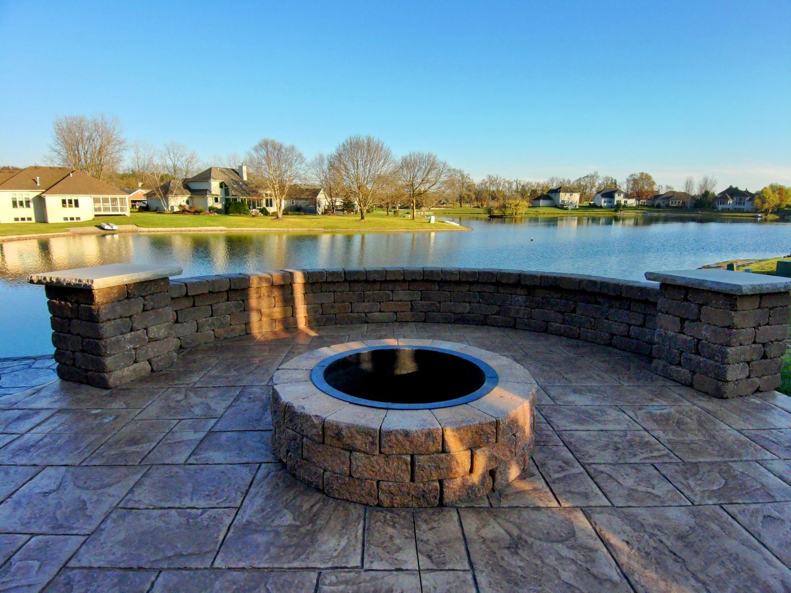 Lakeside stamped concrete patio featuring a circular stone fire pit and curved retaining wall seating, offering a scenic and serene outdoor gathering space with waterfront views in a suburban neighborhood.