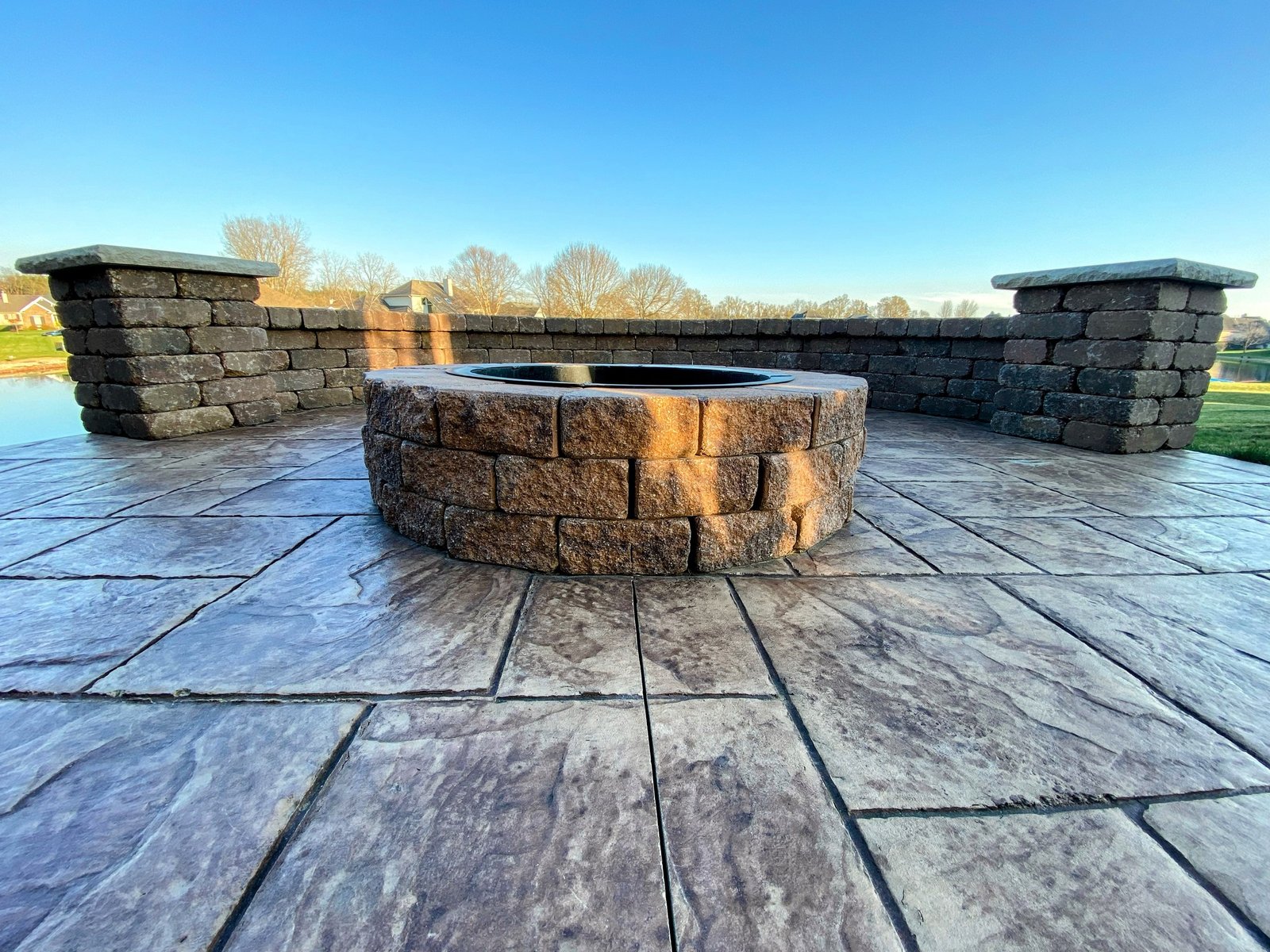 Stamped concrete in Columbia City, Stone fire pit built with stacked bricks, set on a stamped concrete patio with matching stone walls surrounding the space. The background shows bare trees against a clear blue sky, suggesting a crisp outdoor setting. Stamped concrete in Roanoke.