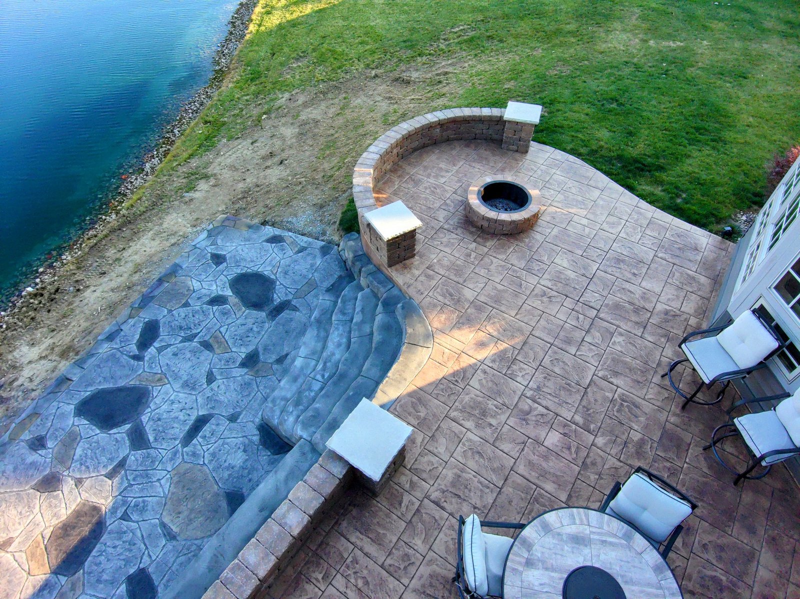 Aerial view of a stamped concrete patio with a built-in fire pit, seating wall, and outdoor furniture overlooking a pond and grassy yard. Stamped concrete in Pierceton