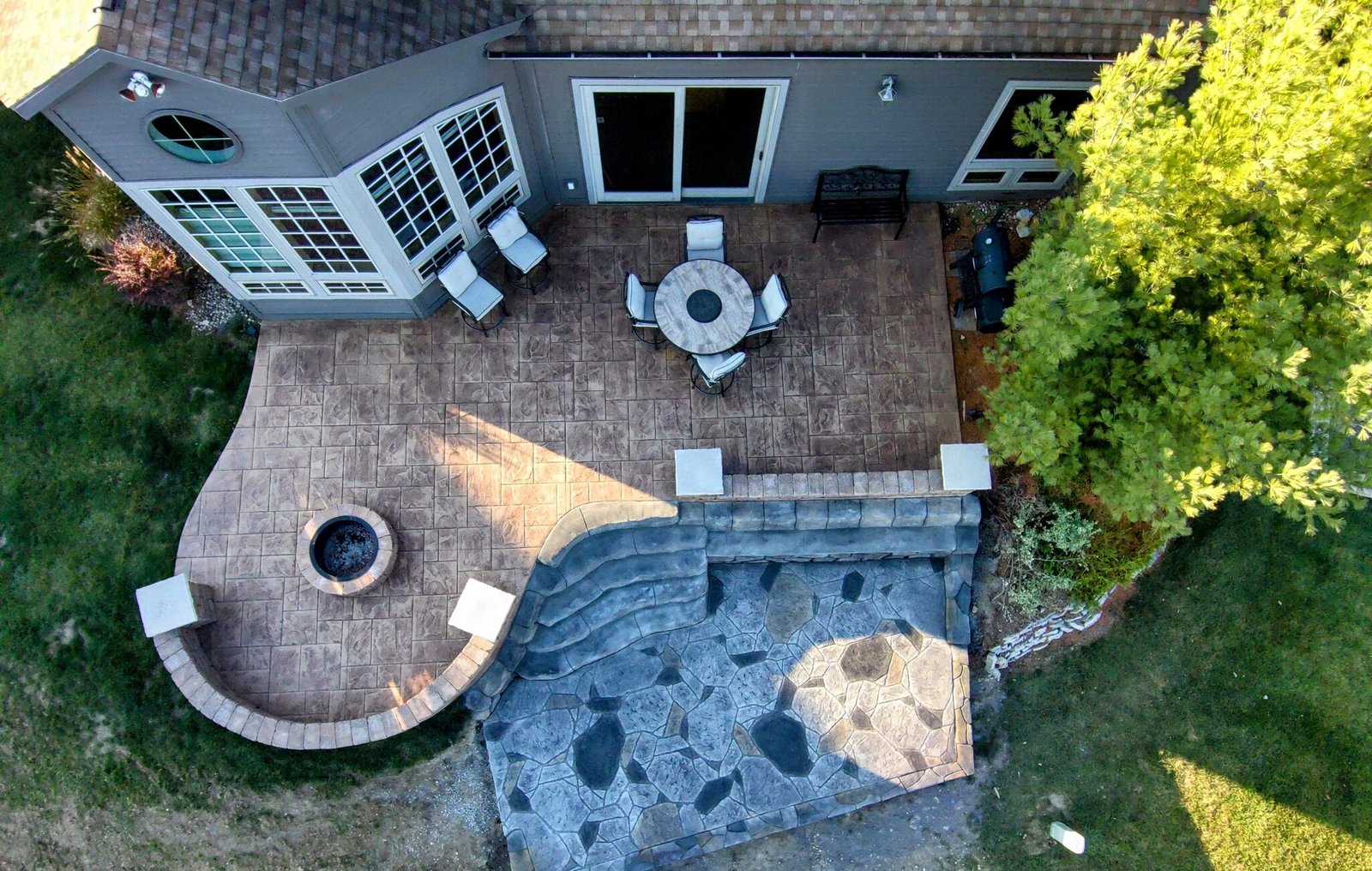Aerial view of a custom multi-level outdoor living space featuring a stamped concrete patio with fire pit and dining area, decorative stone retaining wall, and lower-level flagstone-style concrete pad, seamlessly integrated into a residential backyard landscape.