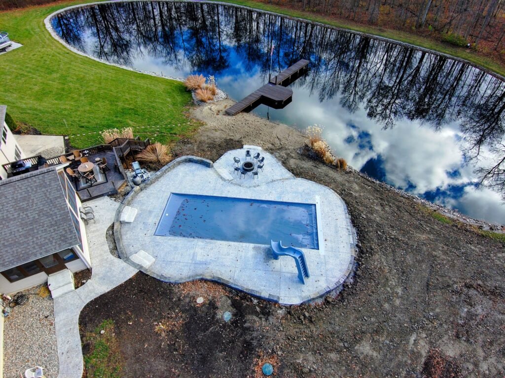 Aerial view of inground swimming pool with stamped concrete deck located next to a pond and landscaped backyard.