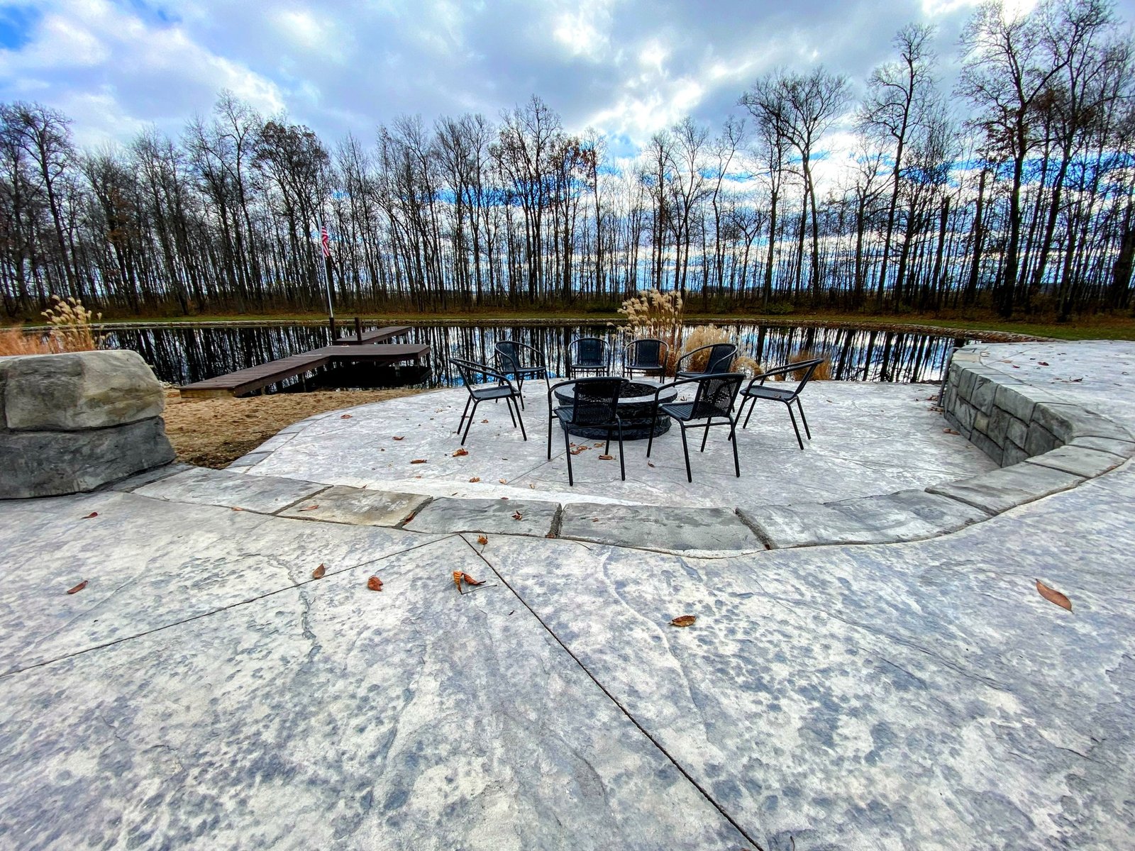 Stamped concrete patio with outdoor seating area beside a pond and wooded backdrop in late autumn. Stamped concrete in Bluffton.