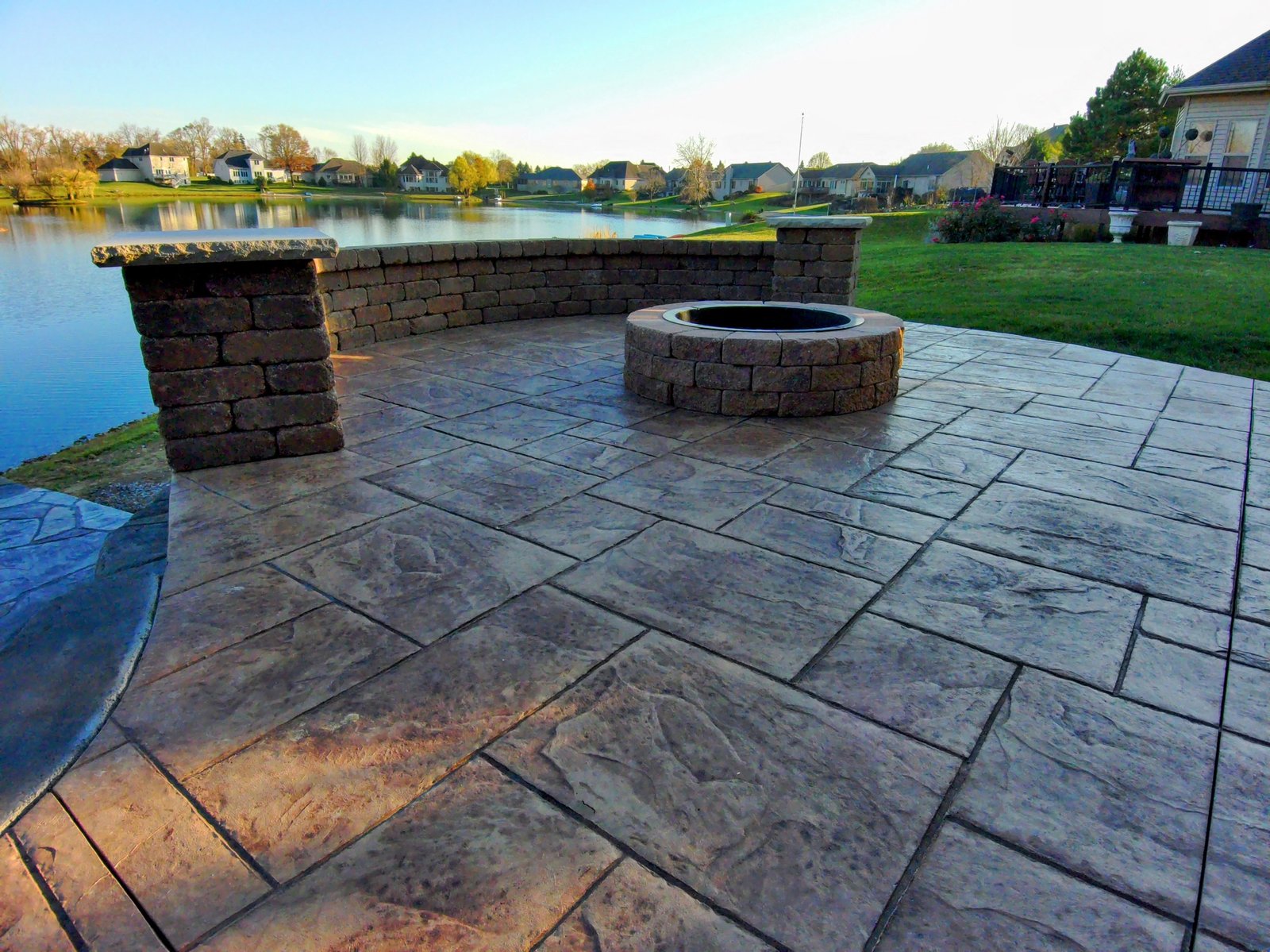 Stamped concrete patio with stone fire pit and curved seating wall overlooking a backyard pond and neighborhood homes. Concrete patios in Churubusco. Stamped concrete in Huntertown. Concrete contractor in Goshen