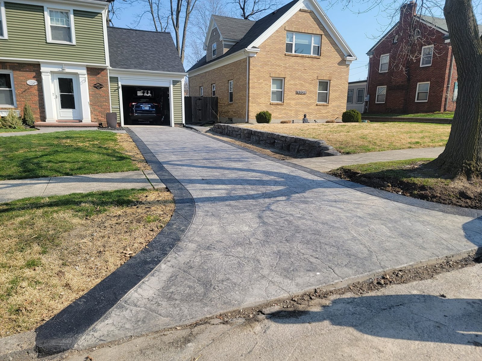 Stamped concrete driveway leading to single-car garage of a brick home in a suburban neighborhood. Driveway replacement in Warsaw. Driveway replacement in Huntington. Driveway replacement in Bryan