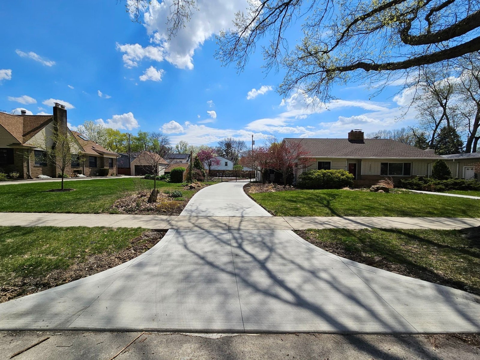 Curved light gray concrete driveway leading to a residential home with landscaped yard and spring trees under a blue sky. Driveway replacement in North Manchester. Driveway replacement in Avilla. Driveway replacement in Albion. Driveway replacement in Goshen. Driveway replacement in Middlebury