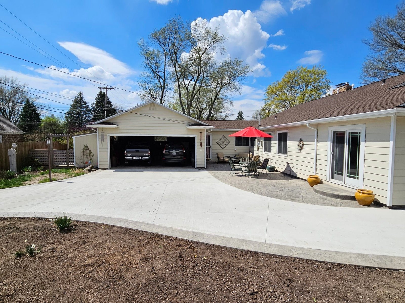 Freshly poured concrete driveway leading to a two-car garage, with an adjacent stamped concrete patio featuring outdoor furniture and a red umbrella in a residential backyard. Driveway replacement in Leo-Cedarville. Driveway replacement in Columbia City. Driveway replacement in Decatur. Concrete contractor in Albion. Driveway replacement in Angola.