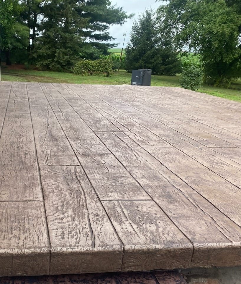 Stamped concrete patio with a wood plank texture finish, installed in a backyard setting surrounded by trees and greenery.