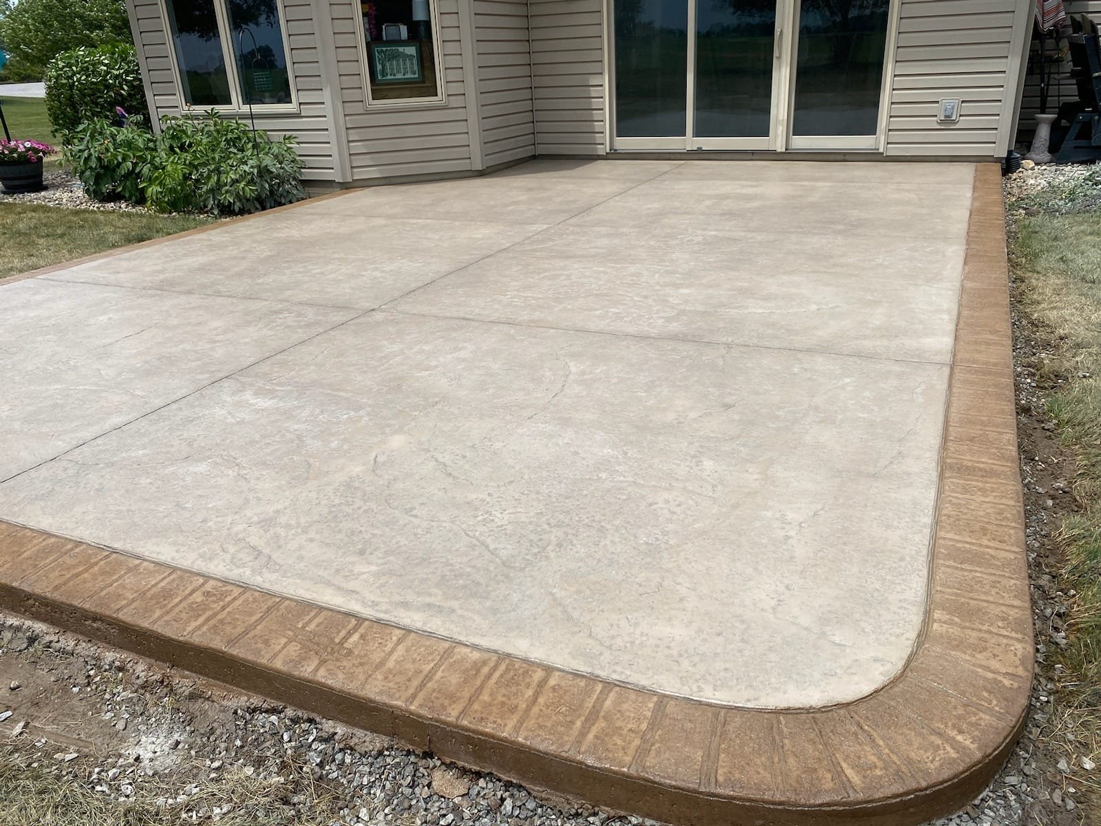 Stamped concrete patio with a smooth, light beige surface and a decorative brown stamped brick border, adjoining a house with beige vinyl siding and sliding glass doors. Stamped concrete in Wabash.