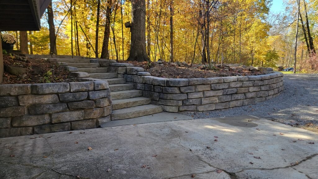 Tiered concrete block retaining wall with integrated steps, blending into a wooded landscape and providing structural support and erosion control on a sloped property.
