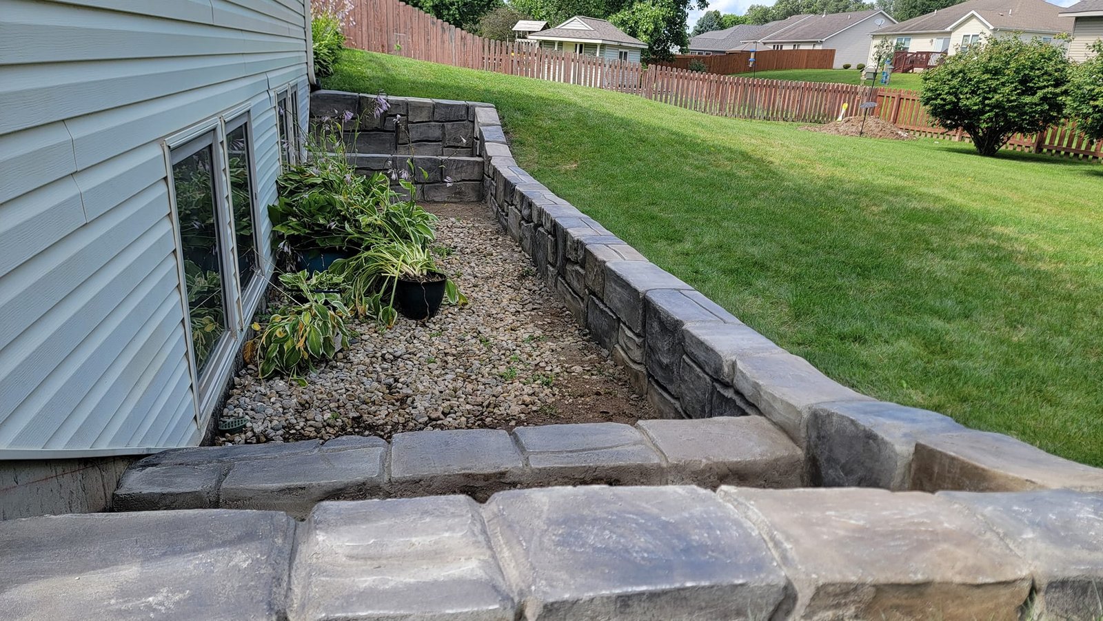 Stone retaining wall surrounding a landscaped garden bed with gravel and plants, set against a sloped grassy yard. Concrete contractor in Warsaw.