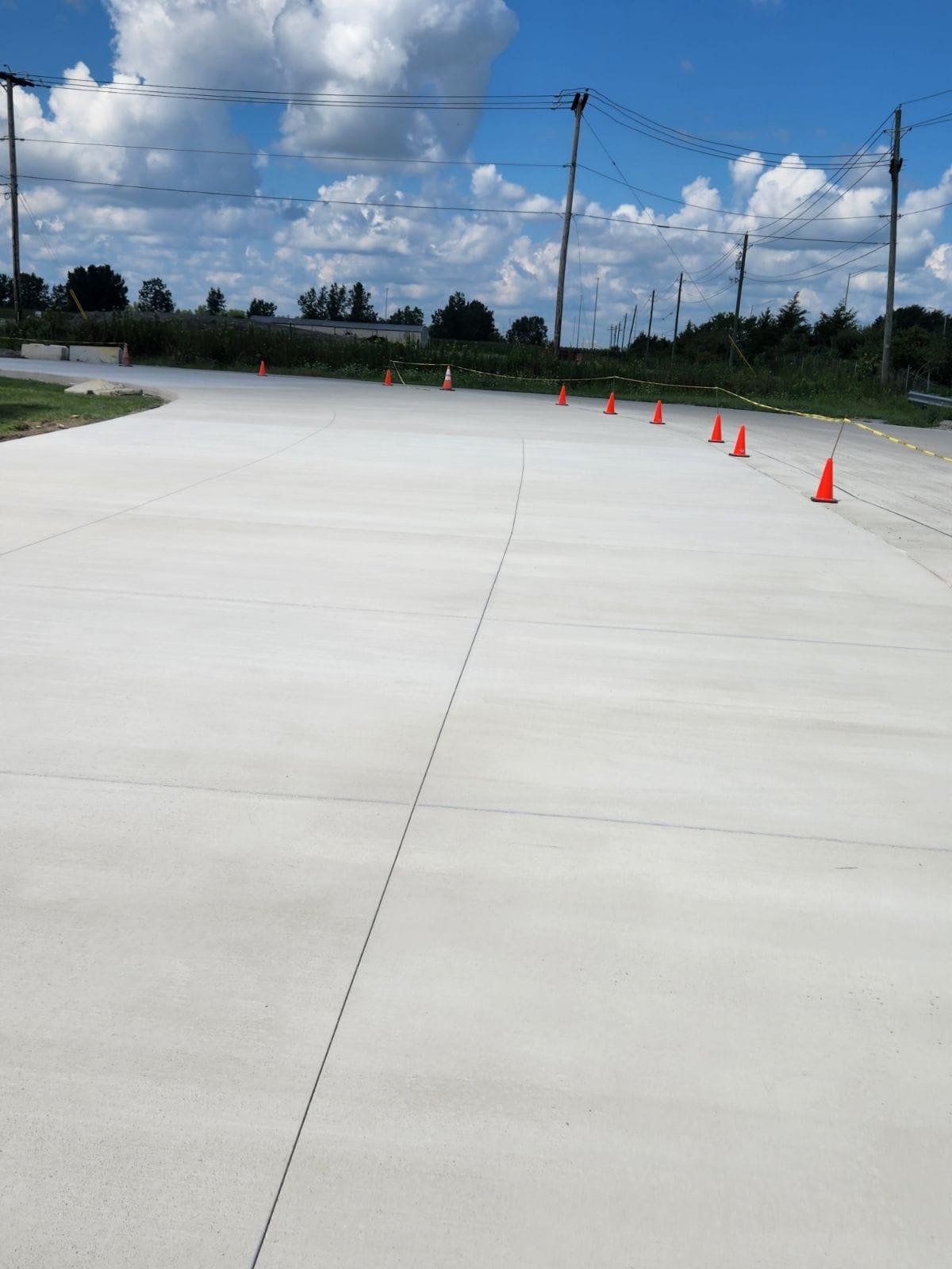Wide expanse of freshly poured concrete pavement with traffic cones along the edge, bordered by grass and utility poles under a partly cloudy sky. Commercial concrete contractor in North Manchester.
