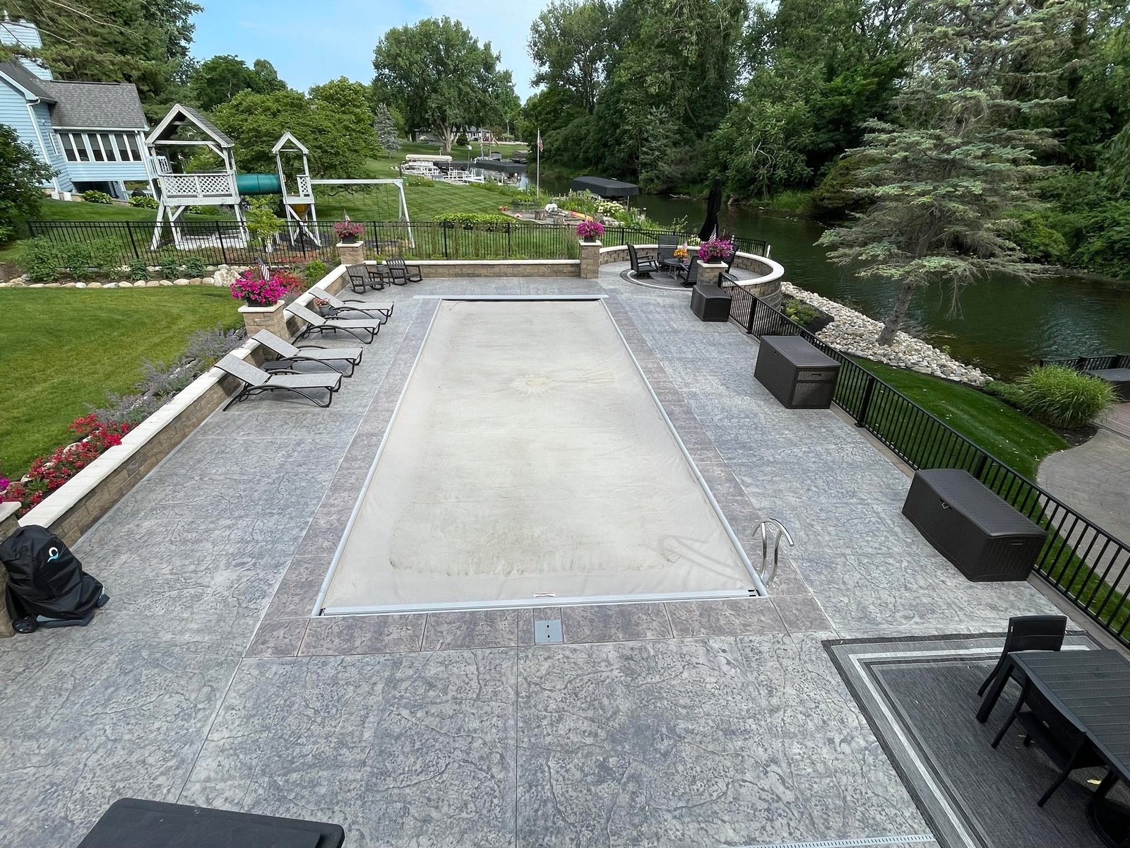 Spacious backyard patio area with lounge chairs, outdoor sofas, and potted flowers, overlooking a landscaped lawn and water feature. Stamped concrete in Montpelier
