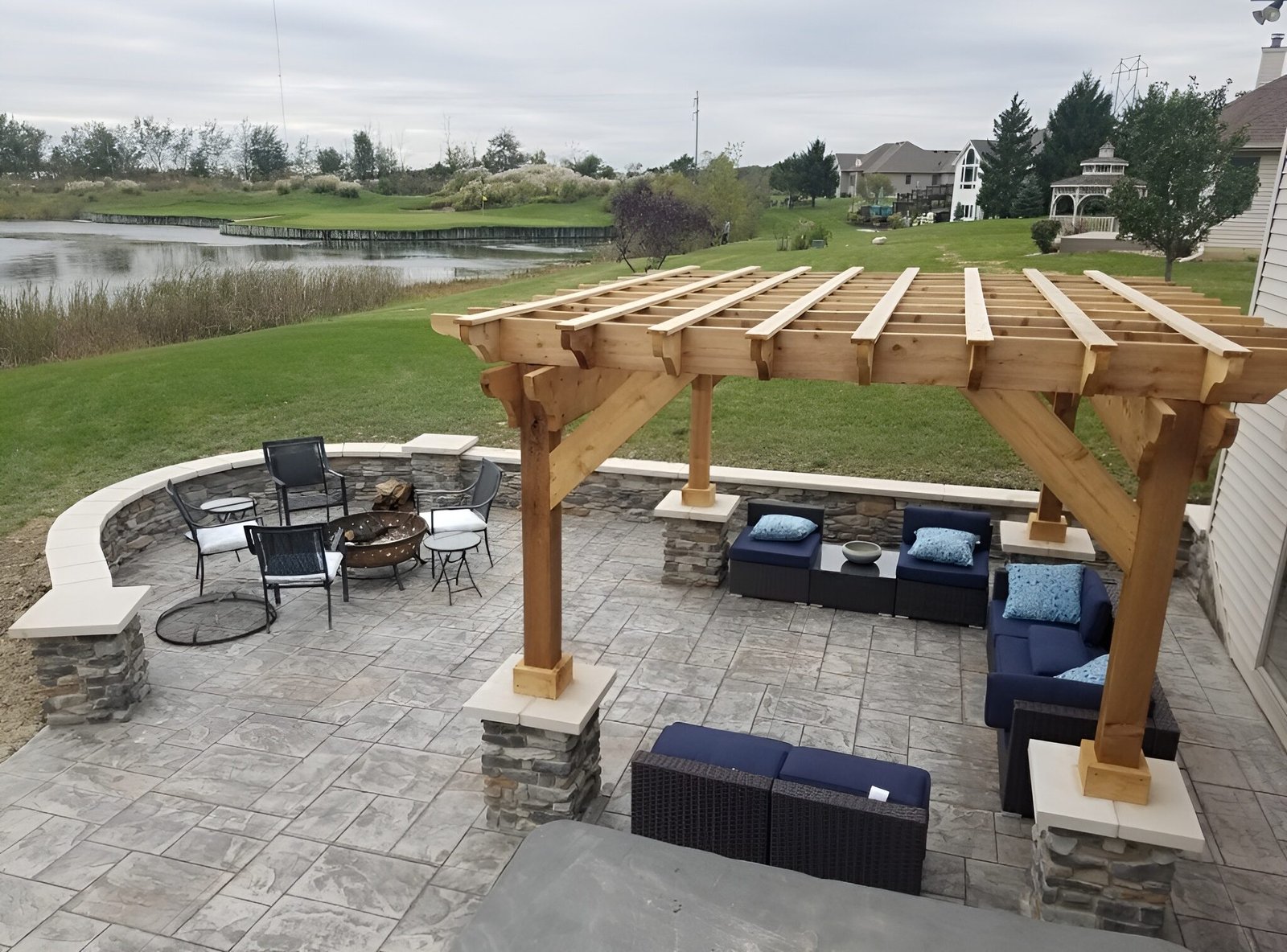 Stamped concrete patio featuring a wooden pergola, curved seating wall, and fire pit overlooking a scenic pond. Concrete contractor in Auburn. Concrete contractor in Defiance