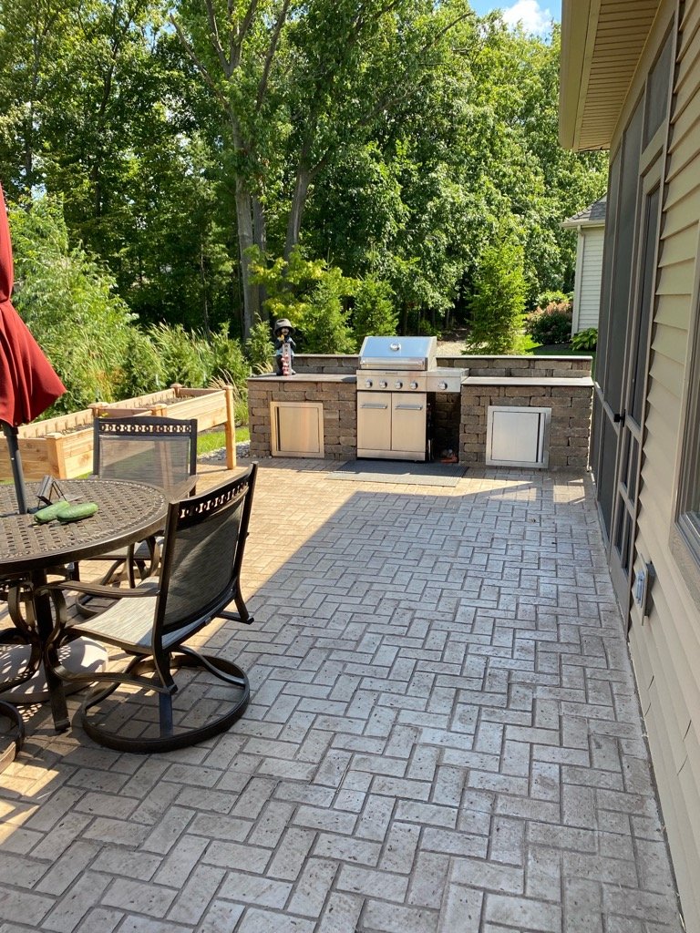 Stamped concrete patio with a herringbone pattern finish, featuring a built-in outdoor kitchen and grill station with stone veneer and stainless steel cabinets. Concrete patios in Leo-Cedarville
