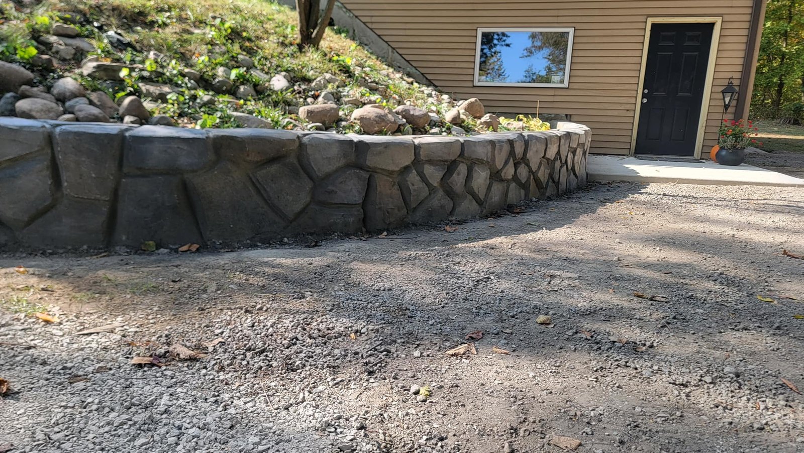 Natural stone retaining wall supporting a sloped garden bed with rocks and greenery, located beside a house with tan siding.”