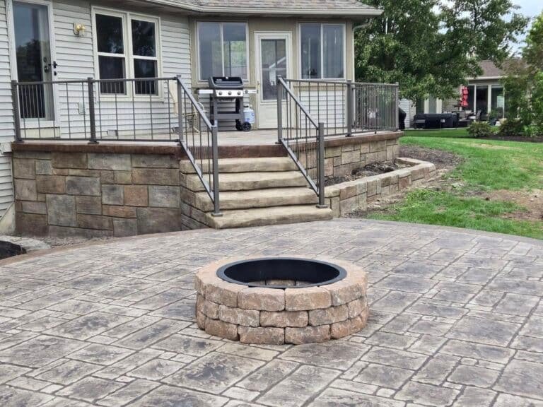 Stamped concrete patio with a built-in stone fire pit and steps leading to a raised deck area.