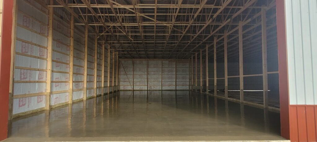 Large indoor concrete slab foundation freshly poured inside a framed commercial or agricultural structure, ready for further construction and load-bearing use.