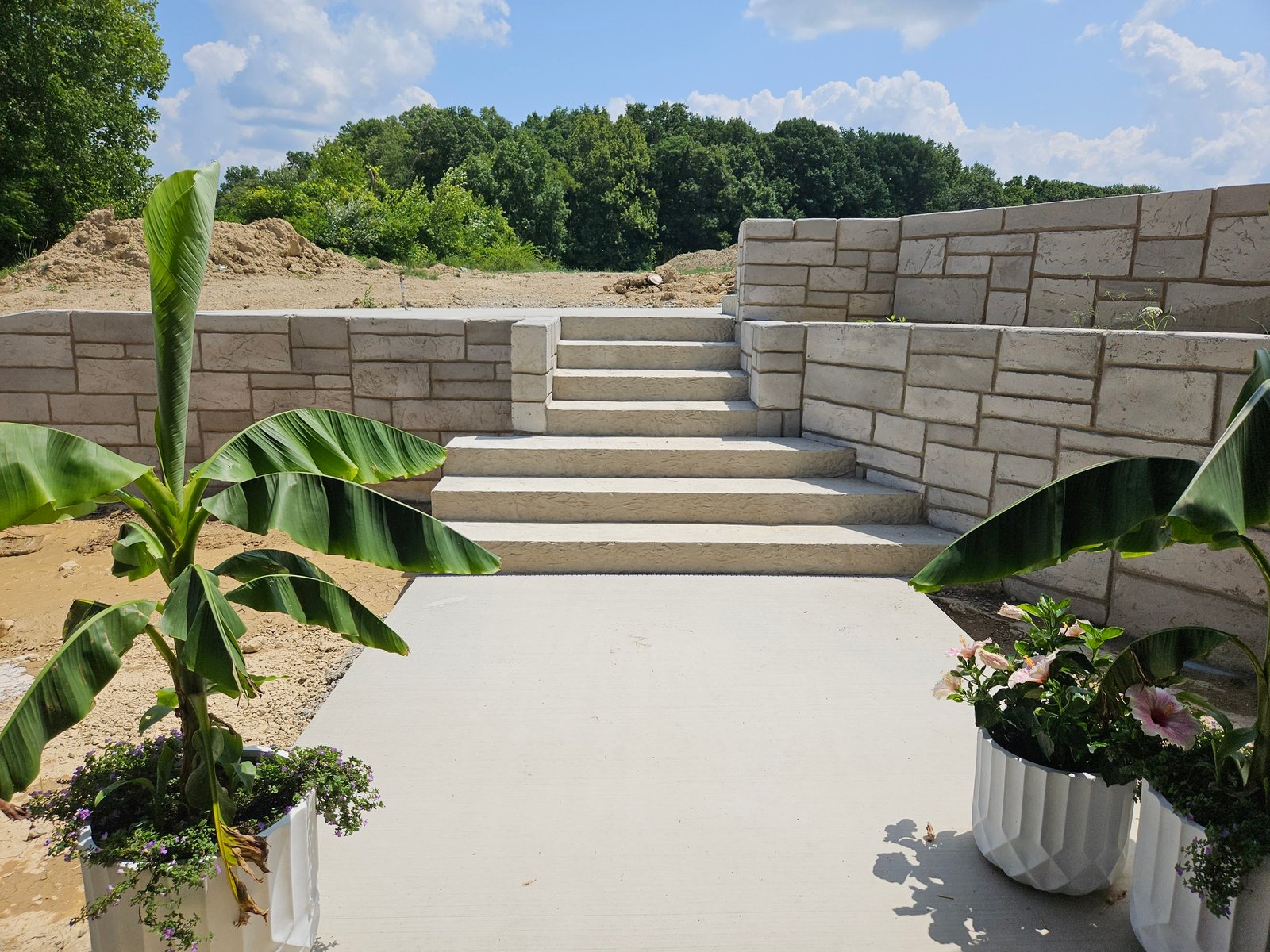 Freshly poured concrete patio with stair access framed by retaining walls and potted plants in a landscaped outdoor area. Concrete contractor in New Paris