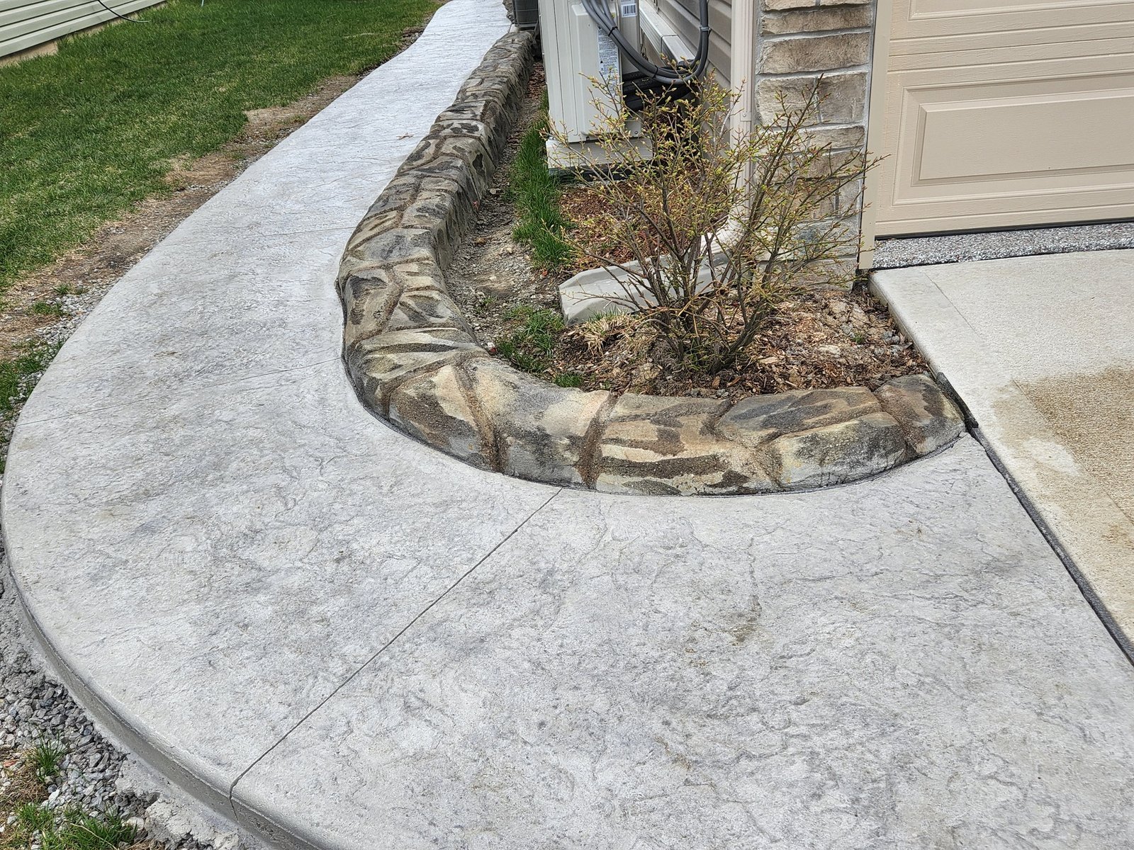 Stamped concrete in Leo-Cedarville. walkway with decorative concrete curbing surrounding a small landscaped area next to a home, showcasing custom stone-textured edging by a concrete contractor.