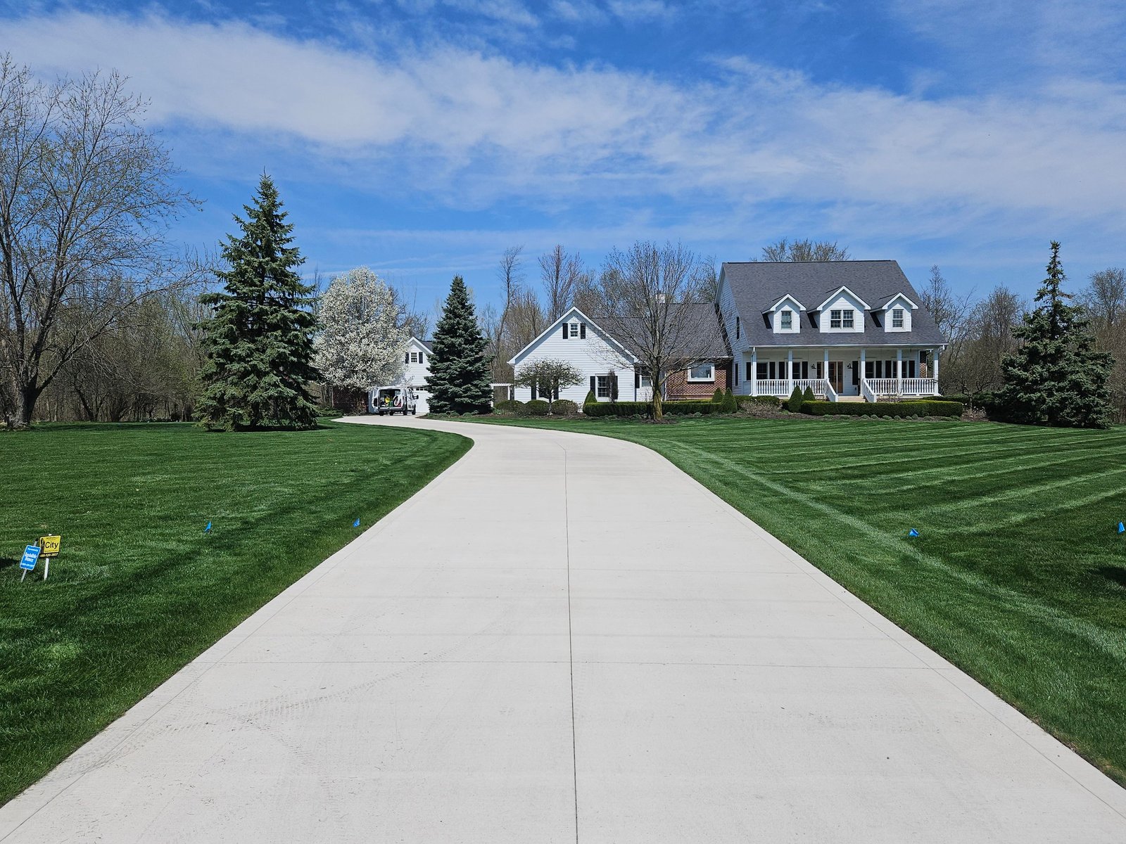 Long concrete driveway leading to a white two-story house with dormer windows, surrounded by green grass, trees, and a bright blue sky with light clouds. Driveway replacement in Berne. Driveway replacement in Butler. Driveway replacement in Ligonier