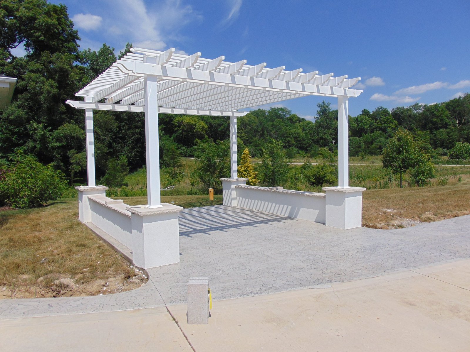 A white pergola with a slatted roof stands on a stamped concrete patio, flanked by short white stucco walls, set against a backdrop of greenery and a bright blue sky. Concrete Contractor in Berne. Concrete contractor in LaGrange