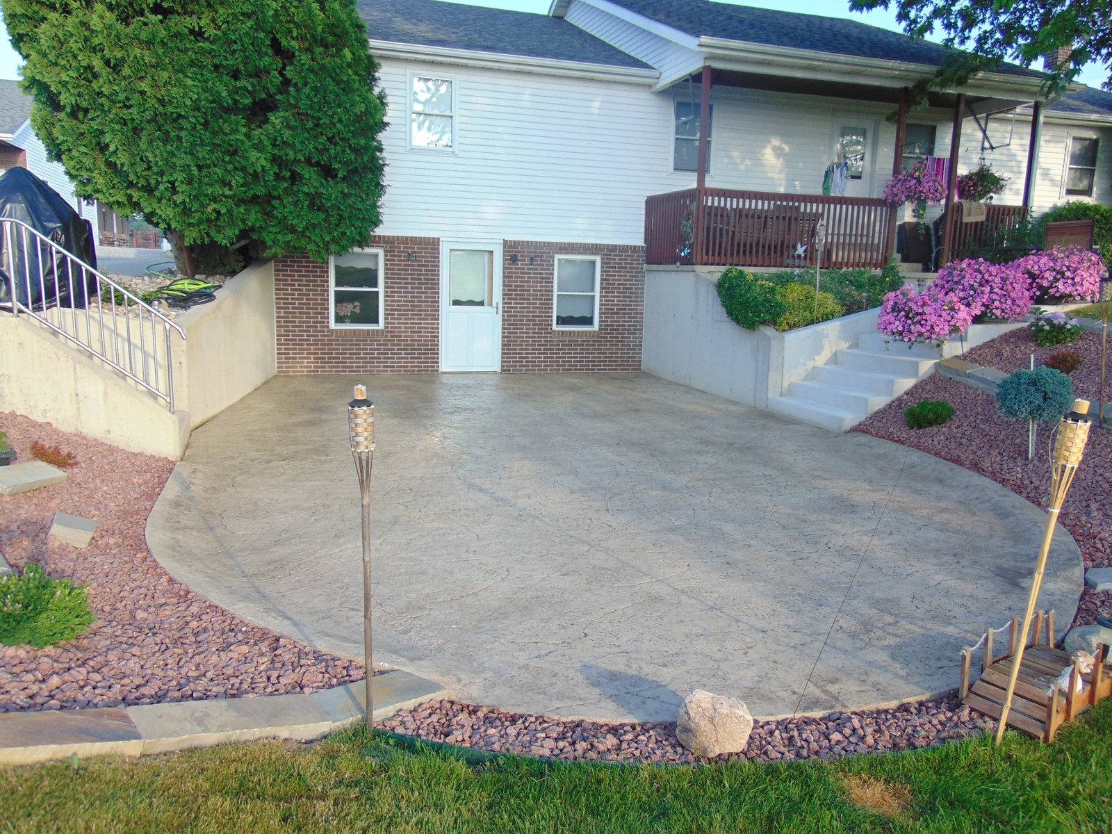 Concrete patios in South Whitley. Stamped concrete patio in front of a split-level house with white siding and brick accents. The patio is bordered by decorative rock landscaping, a concrete stairway leading to a porch with railings, and lush greenery and flowers. Stamped concrete in Angola. Stamped concrete in Hicksville
