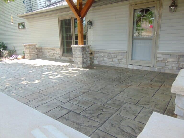 Light gray stamped concrete patio with an ashlar slate pattern, bordered by stone pillars and a house with light siding and stone veneer. Concrete patios in New Paris. Stamped concrete in Defiance