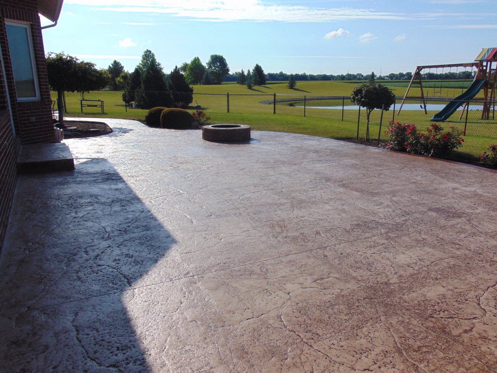Large stamped concrete patio with a circular stone fire pit, overlooking a landscaped backyard with manicured bushes, open green fields, and a fenced pond under a bright blue sky. Concrete Contractor in North Manchester. Concrete contractor in Angola.