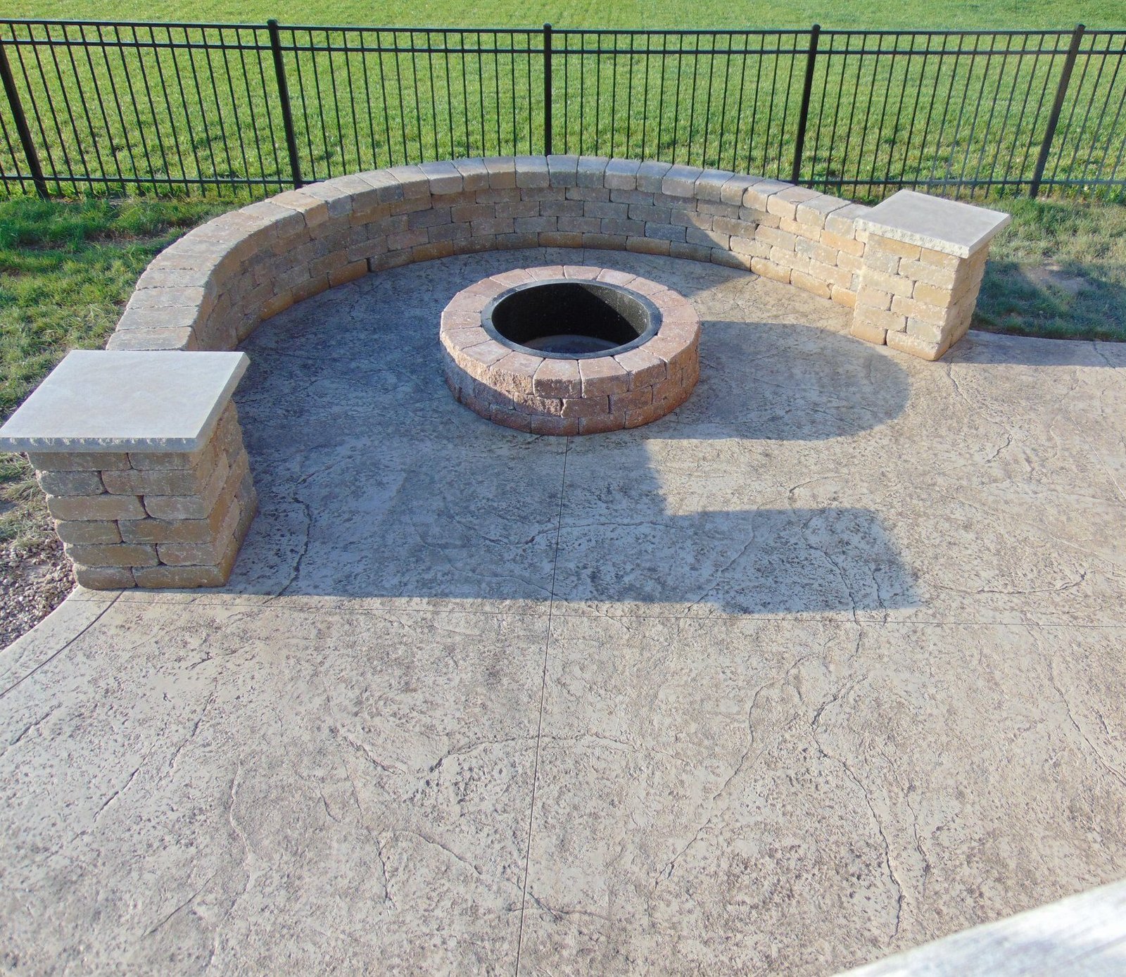 Stamped concrete patio with a built-in stone fire pit and curved block seating wall, enclosed by a black metal fence and surrounded by open green lawn—ideal for backyard entertaining. Stamped concrete in Avilla.