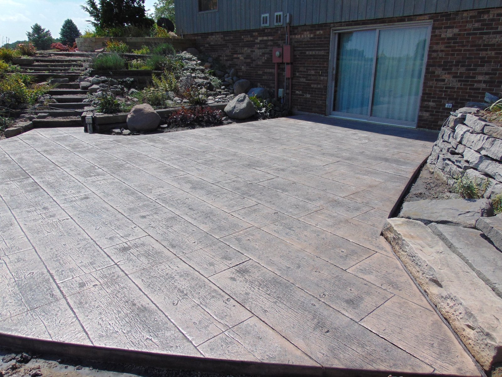 Stamped concrete patio with wood-grain texture beside a brick house and landscaped rock garden. Stamped concrete in North Manchester. Stamped concrete in Paulding.
