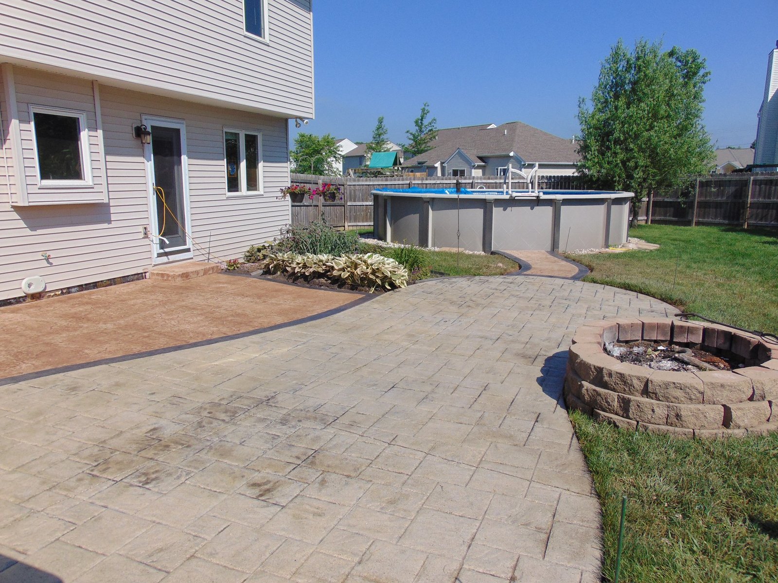 Backyard with a paved stone patio, circular fire pit, and above-ground pool with surrounding deck, adjacent to a beige house with white trim and a garden bed. Concrete patios in Roanoke. Concrete patios in Berne.