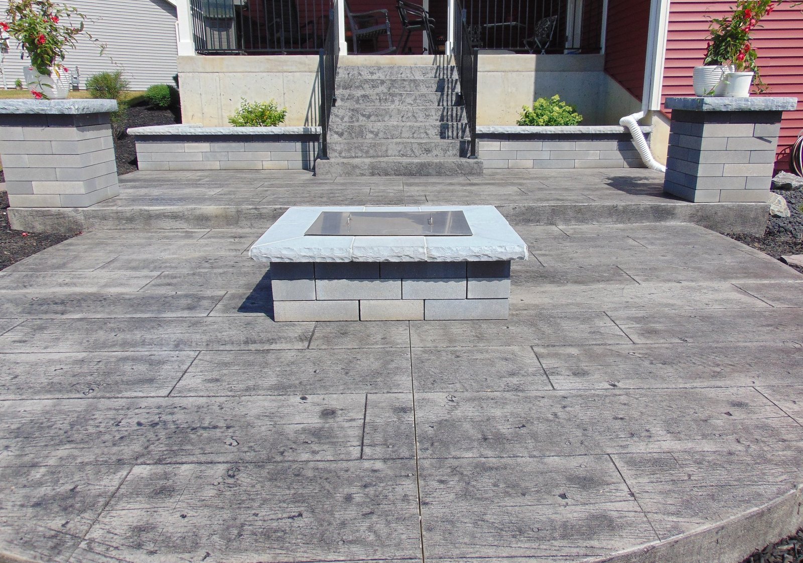 Modern backyard patio with a square stone fire pit in the center, featuring textured concrete flooring and steps leading up to a raised seating area with railings.