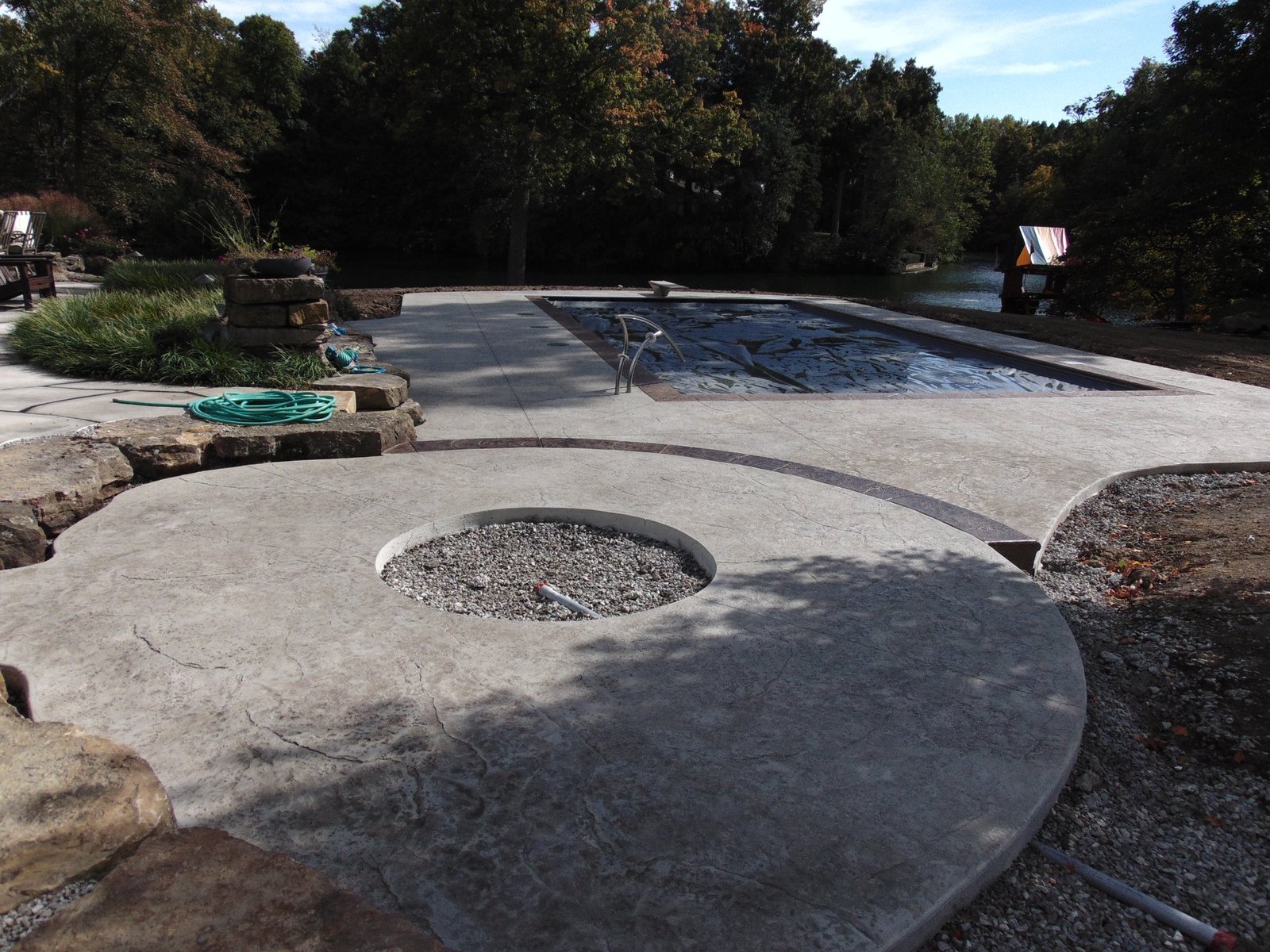Outdoor pool area with a circular concrete fire pit surrounded by gravel, curved concrete patio sections, and wooded landscaping in the background. Stamped concrete in Waterloo