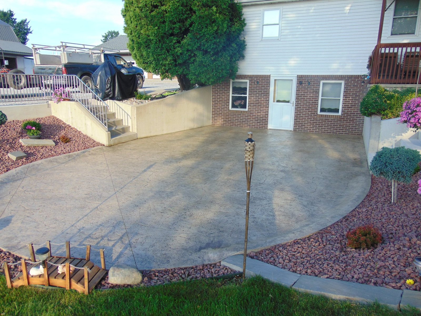 Residential backyard with a smooth concrete patio bordered by decorative rock landscaping and leading to a brick house with basement windows. Concrete patios in North Manchester.