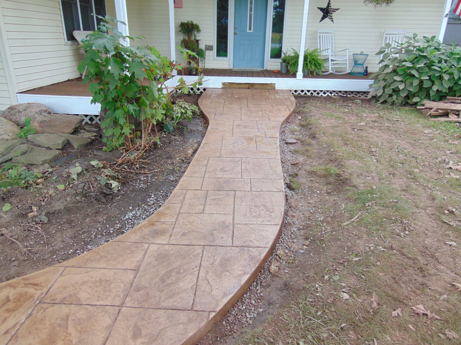 Curved stamped concrete walkway with a decorative square tile pattern leading to a light-colored house with a blue front door and small porch. Plants border the walkway, and soil surrounds the new installation.