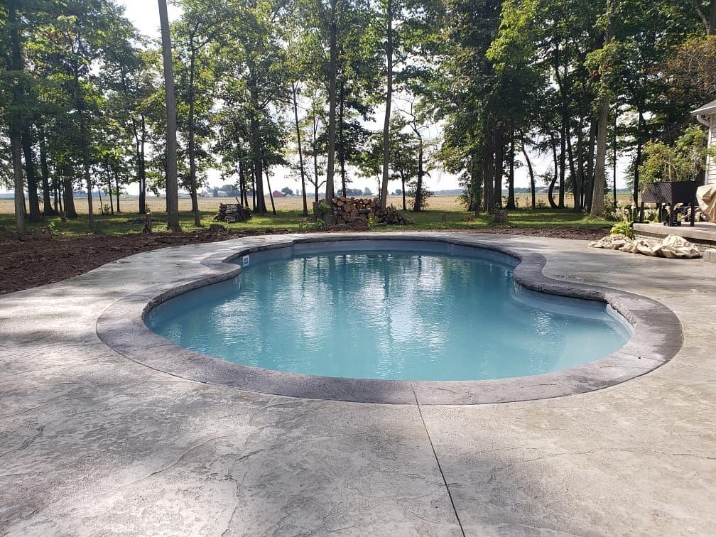 Curved inground swimming pool surrounded by a concrete deck, set in a wooded backyard with tall trees and a view of open land or water in the distance. Stamped concrete in South Whitley. Concrete patios in Butler. Stamped concrete in Kendallville