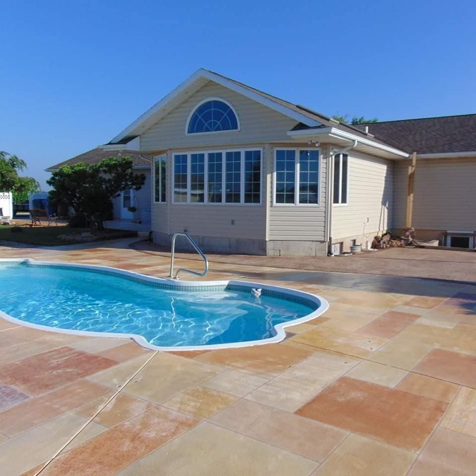 concrete patios in Columbia City. Residential backyard with an inground swimming pool surrounded by multicolored stone pavers. The pool is located beside a light-colored house with large windows and a gable roof, under a clear blue sky. Concrete patios in Ligonier.