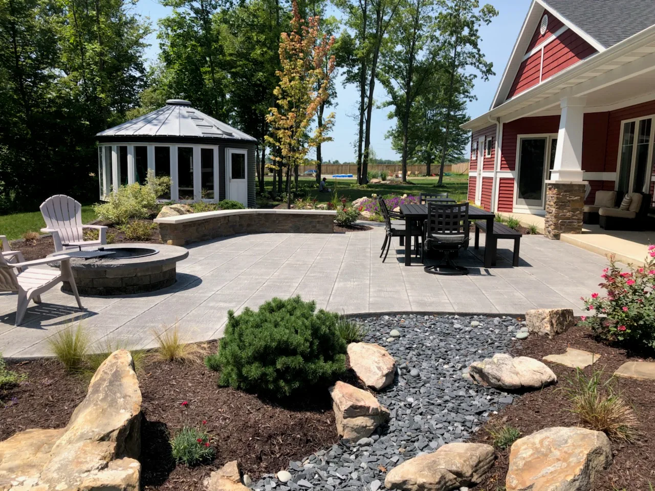 Concrete contractor in Fort Wayne. Spacious backyard patio with clean-cut concrete pavers, built-in stone fire pit, dining area, curved seating wall, and surrounding landscape featuring boulders, mulch, and decorative rock bed, creating a stylish and functional outdoor living space beside a modern farmhouse-style home.