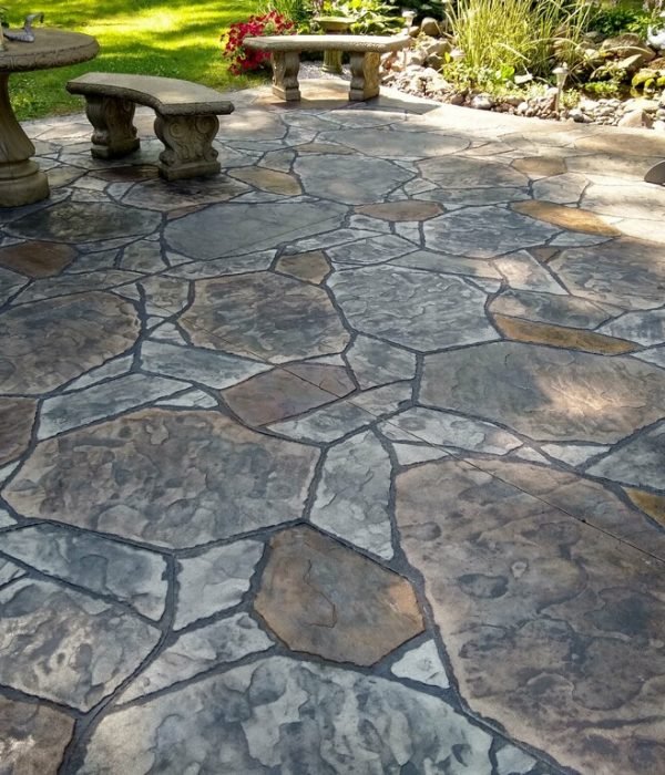 Our services. Outdoor stamped concrete patio with a natural stone pattern and earthy color tones, surrounded by a garden landscape and stone seating, showcasing decorative concrete craftsmanship.