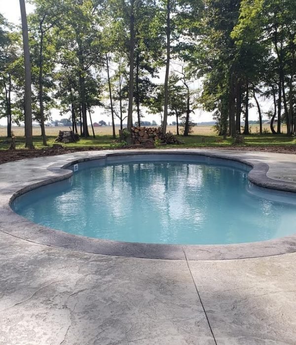 Round concrete pool surrounded by stamped decking and shaded by tall trees, providing a peaceful forest-side retreat.